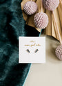 Offset Trio - Sapphire: small blue and gold-plated stud earrings with sapphire CZ stones, displayed on a white JaxKelly “wear good vibes” card, set on teal velvet with pink pom-poms and a beige fan in the background.