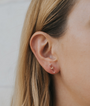 Close-up of a woman's ear with straight blonde hair, wearing the Clear Quartz Mineral Point - Cleansing earring, a minimalist clear stud with 18k gold plating. The softly blurred background enhances the elegant design.