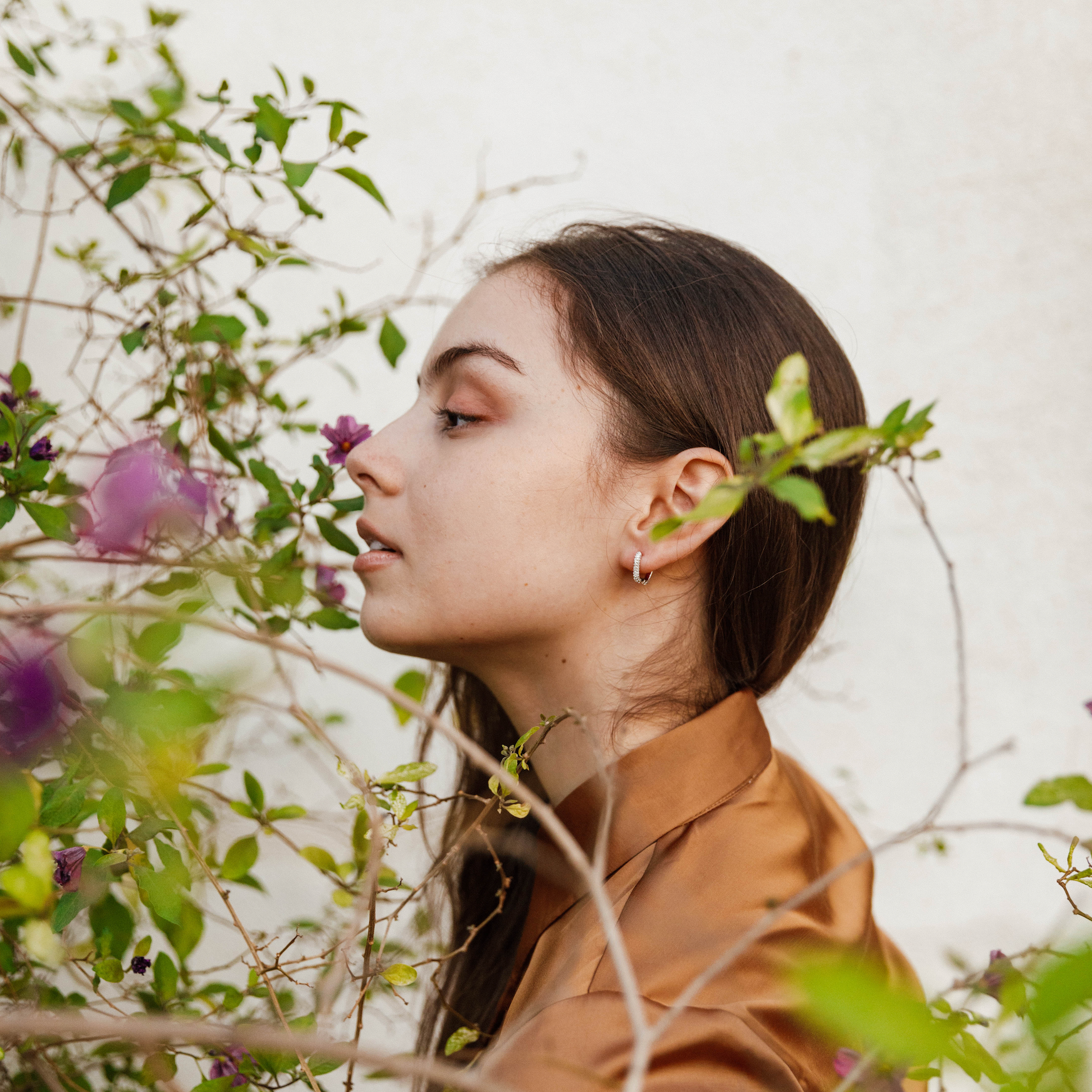 A young woman with long brown hair and a brown shirt stands in profile near green leaves and purple flowers, admiring a bloom. She wears Pavé Silver Hoop earrings, which add a subtle sparkle against the light background.