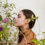 A young woman with long brown hair and a brown shirt stands in profile near green leaves and purple flowers, admiring a bloom. She wears Pavé Silver Hoop earrings, which add a subtle sparkle against the light background.