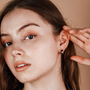 A young woman with long brown hair touches her ear, displaying the Silver Wide Hoop earring with sterling silver posts. She wears natural makeup and stands against a neutral background, reflecting minimalist style.