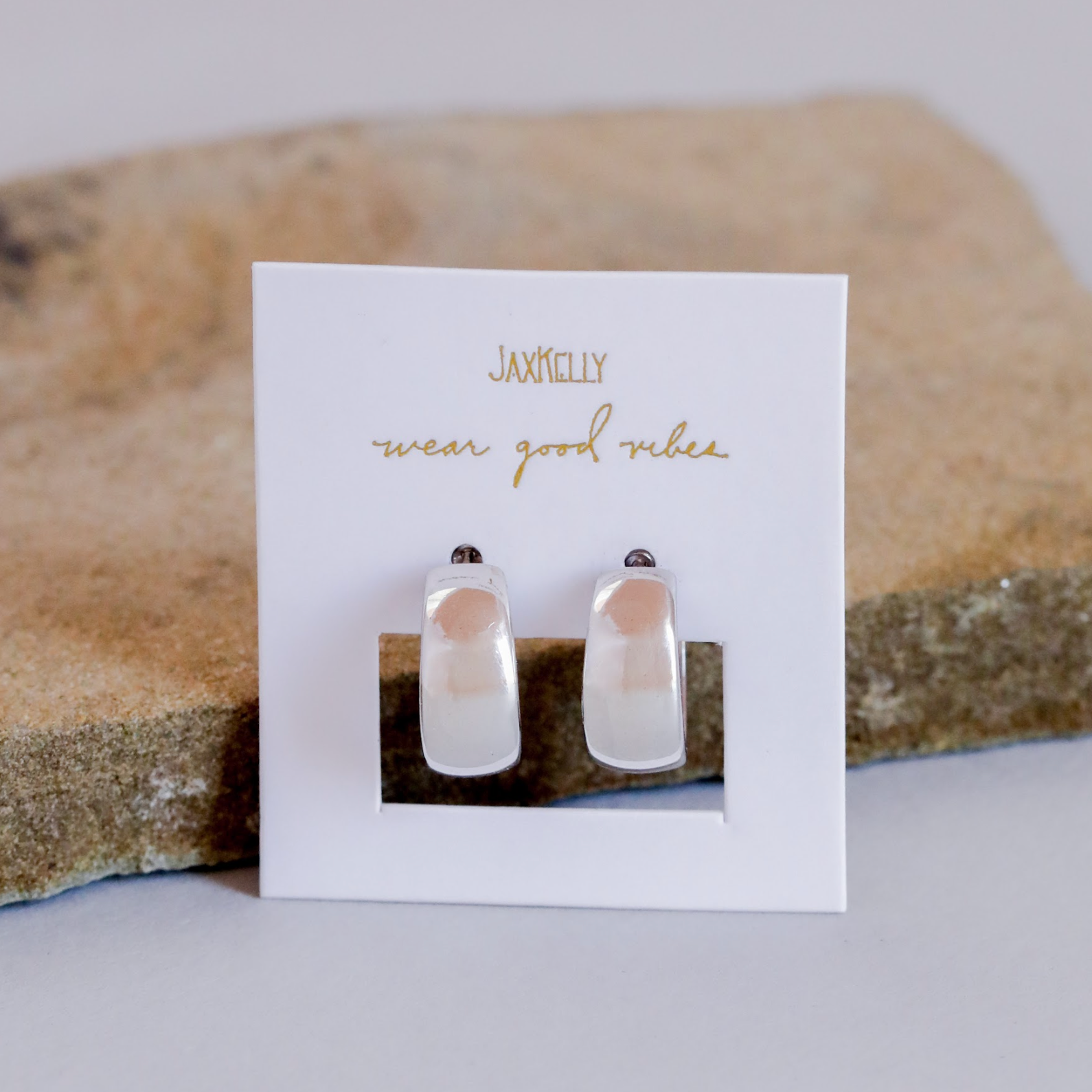 A pair of Silver Wide Hoops with a modern minimalist design and sterling silver posts, displayed on a white JaxKelly “wear good vibes” card against a neutral stone background.
