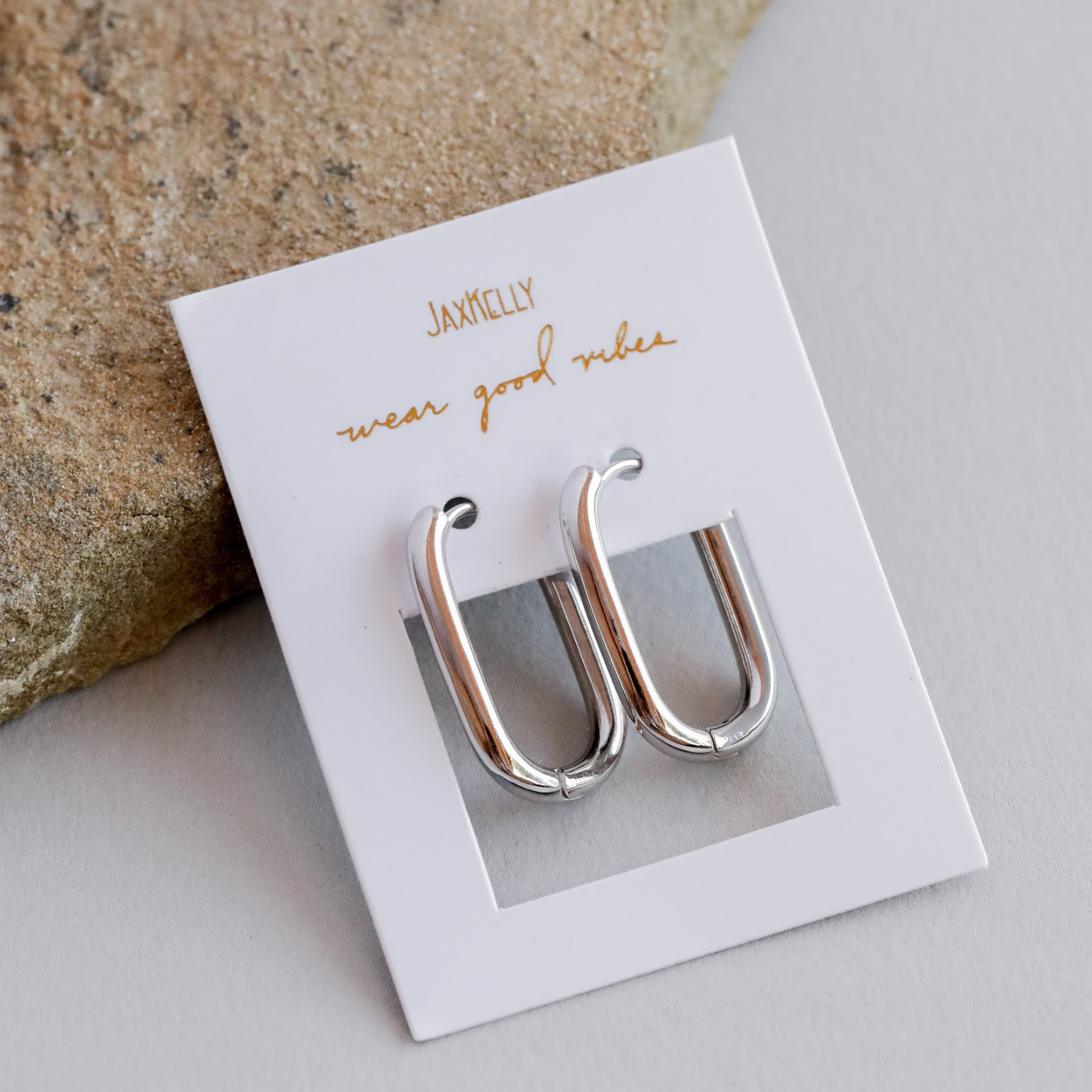 The Silver Rectangle Hoop - Large earrings are displayed on a white jewelry card with gold “JaxKelly wear good vibes” text, next to beige stone on a light gray surface.
