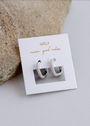 A pair of Pavé Silver Hoop earrings encrusted with small crystals sits on a white JaxKelly card reading "wear good vibes," placed on a light gray surface next to a textured stone.