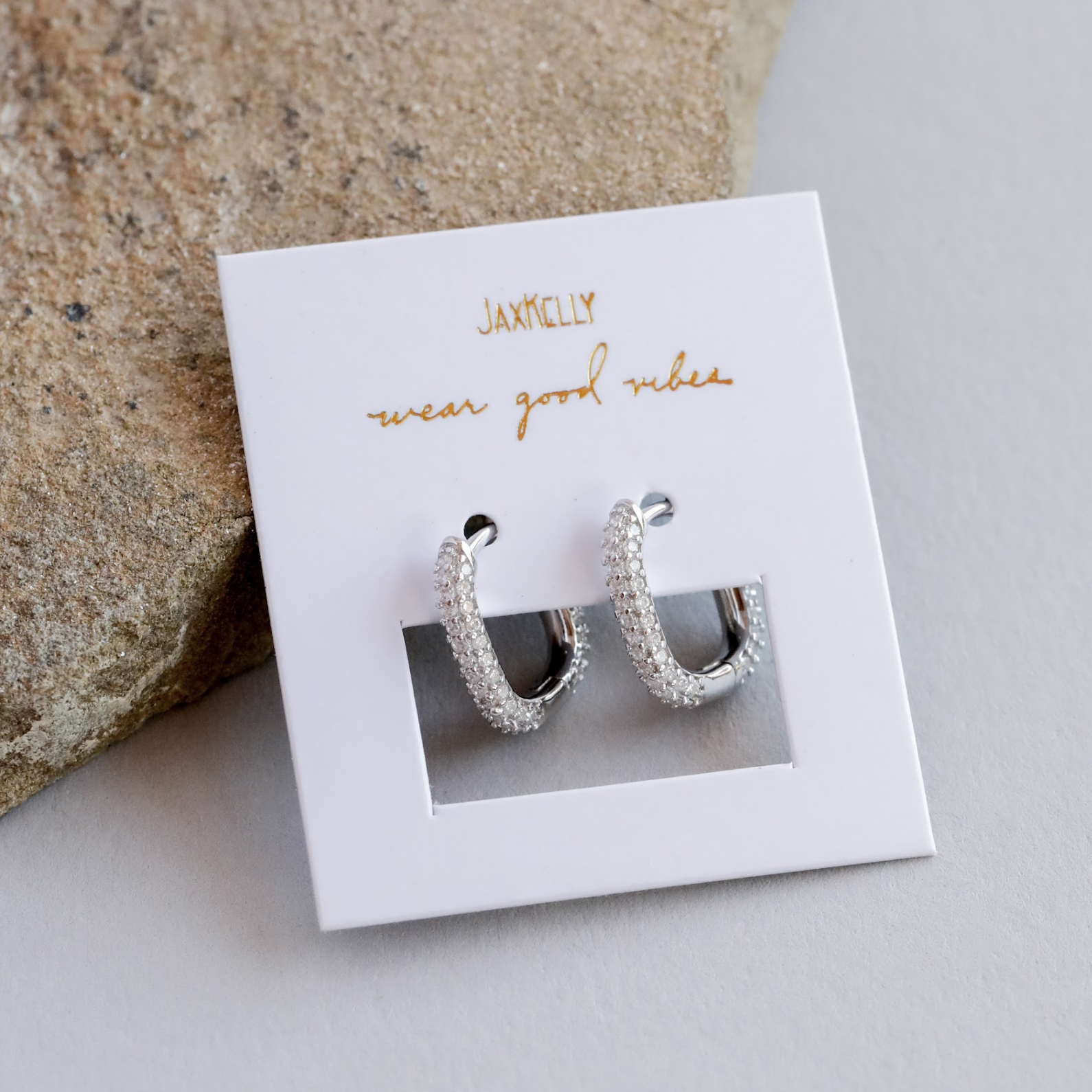 A pair of Pavé Silver Hoop earrings encrusted with small crystals sits on a white JaxKelly card reading "wear good vibes," placed on a light gray surface next to a textured stone.