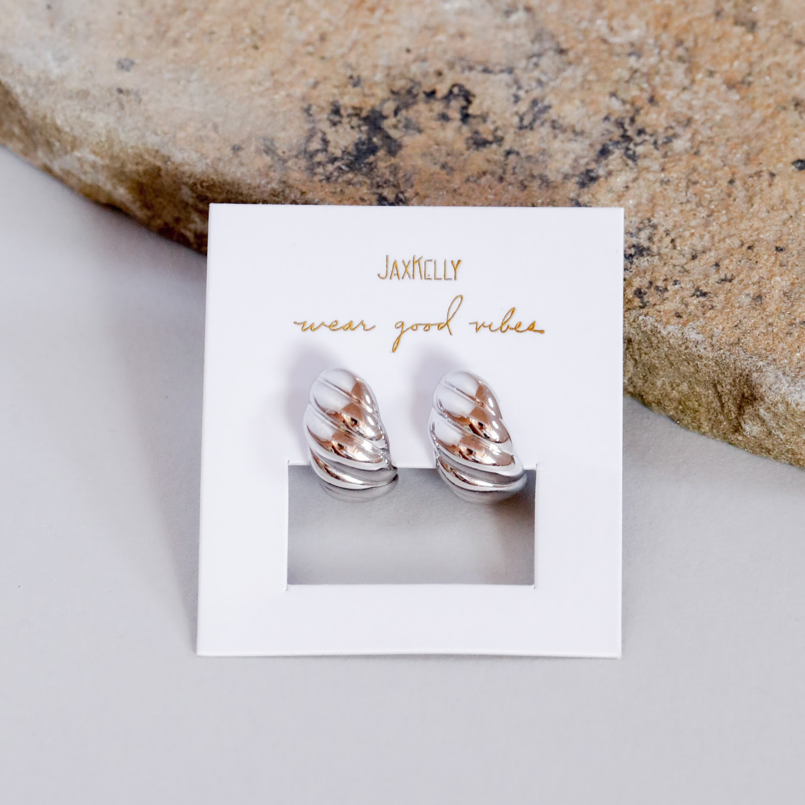 The Silver Swirl Hoop, featuring teardrop-shaped silver and rose gold accents with sterling silver posts, is shown on a white card with gold JAXKELLY wear good vibes text, placed beside a textured stone on a light surface.