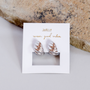 The Silver Swirl Hoop, featuring teardrop-shaped silver and rose gold accents with sterling silver posts, is shown on a white card with gold JAXKELLY wear good vibes text, placed beside a textured stone on a light surface.