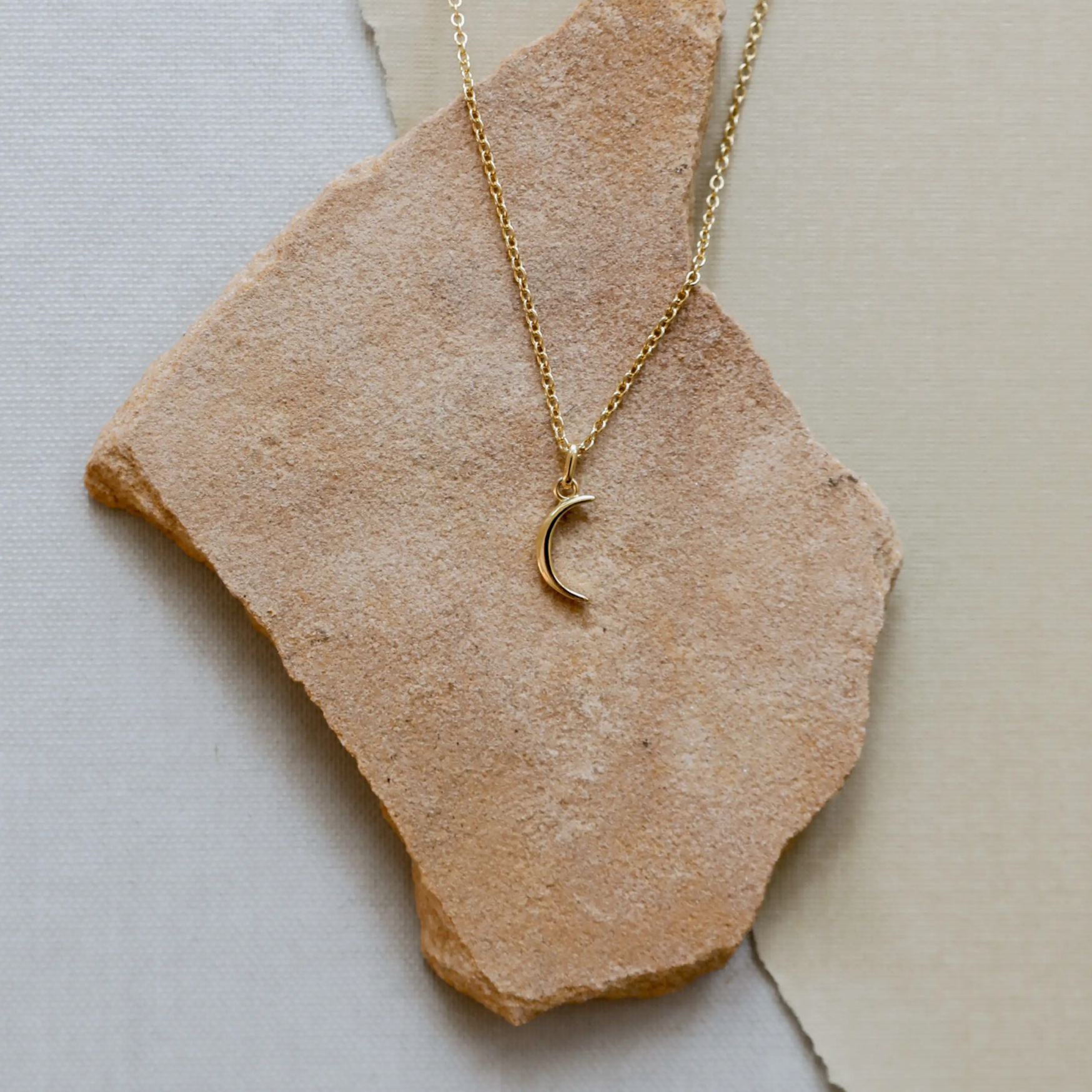 The Tiny Crescent Moon Necklace, crafted with an 18k gold-plated finish and adjustable chain, is showcased on a rough light brown stone against a neutral textured backdrop.