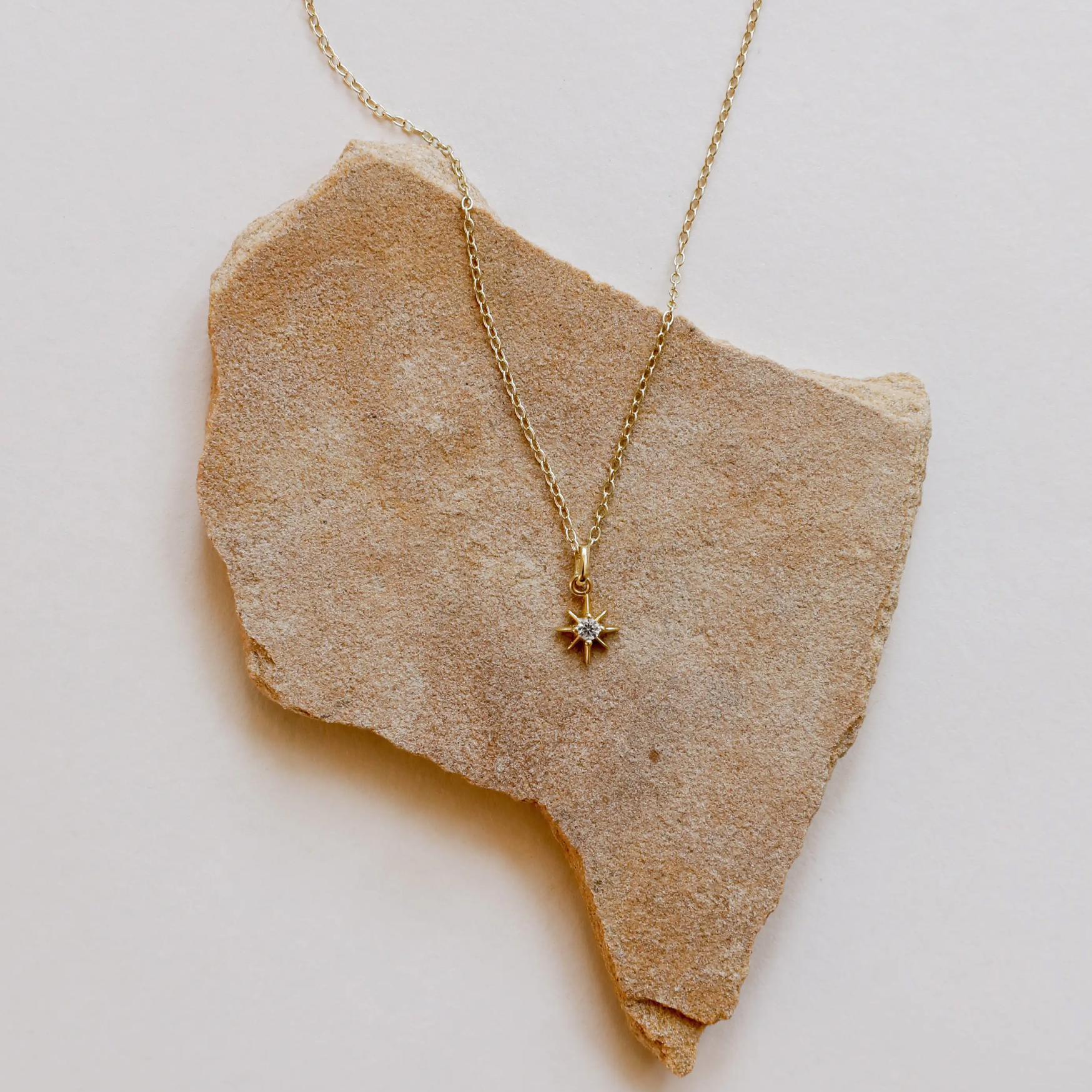 The Tiny Star Necklace, featuring a delicate star-shaped Cubic Zirconia charm, is shown on a rough beige stone slab against a light background.