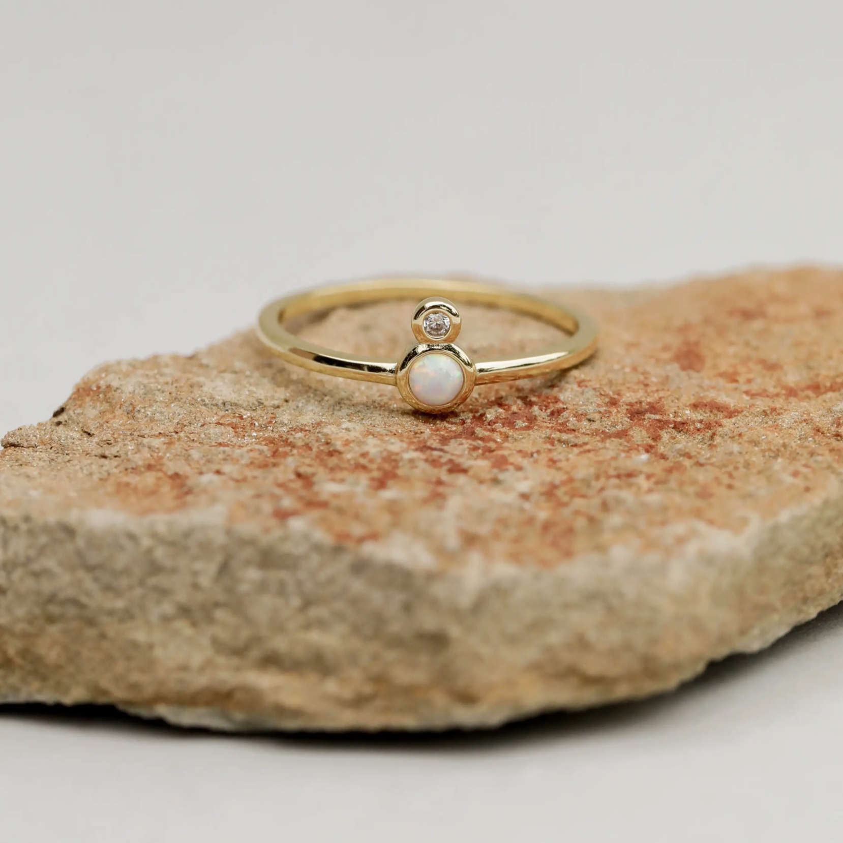 The Opal Infinity Ring, featuring gold plating, a small white gemstone, and a clear CZ stone, is shown resting on a textured light brown rock with a simple backdrop.
