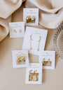Six pairs of Offset Trio - White Opal gold plated earrings, each on a white JaxKelly "wear good vibes" card, displayed on beige fabric near a textured glass. The set features stars, moons, hoops, and lab-grown white opal studs.