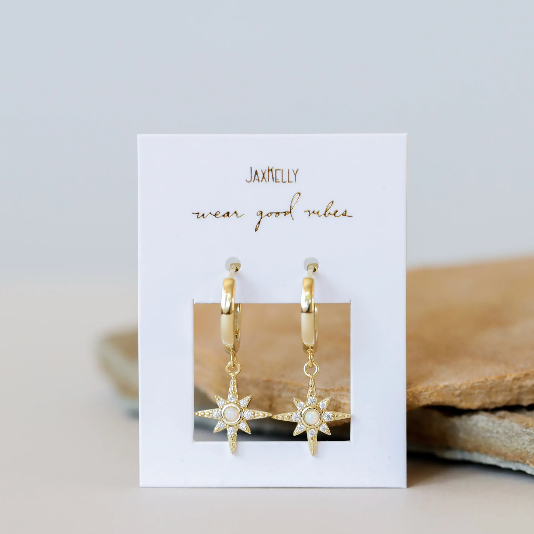 The Starburst Mini Hoop earrings feature gold hoops with starburst charms and small gemstones, displayed on a white card reading "JAXKELLY wear good vibes," against a neutral background.