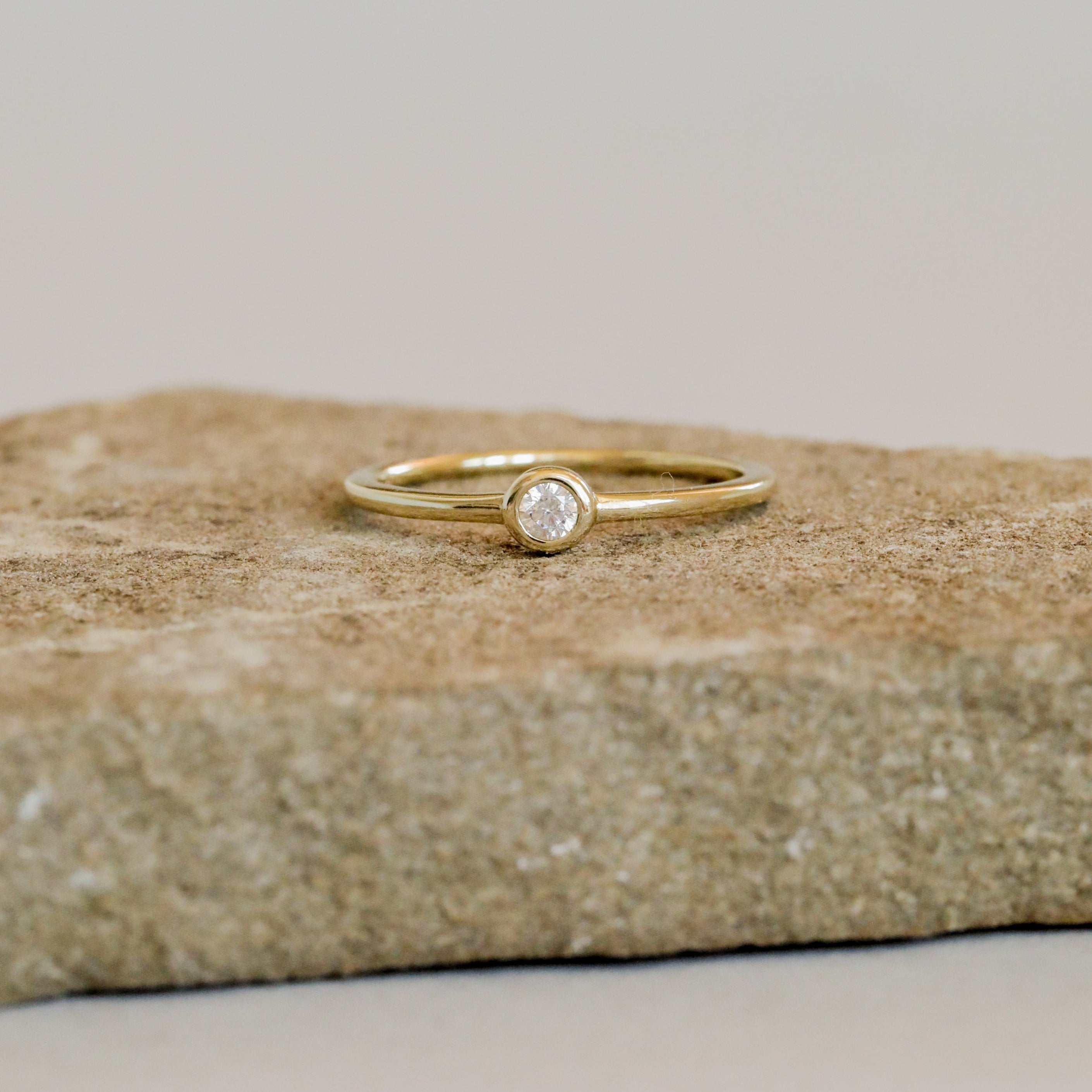 The Simple Stud Ring features a single round diamond in a bezel setting on an 18k gold plated band, shown atop a rough beige stone with a neutral background.