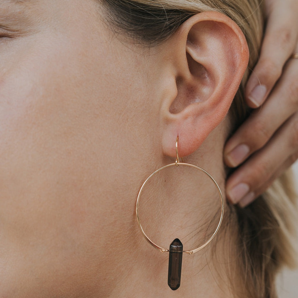 A person wears the Smoky Quartz Hoop - Protection: a large gold hoop earring with a dark smoky quartz crystal pendant. Their hand touches their head, showcasing this handcrafted jewelry; their ear and part of their face are visible.