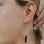 A person wears the Smoky Quartz Hoop - Protection: a large gold hoop earring with a dark smoky quartz crystal pendant. Their hand touches their head, showcasing this handcrafted jewelry; their ear and part of their face are visible.