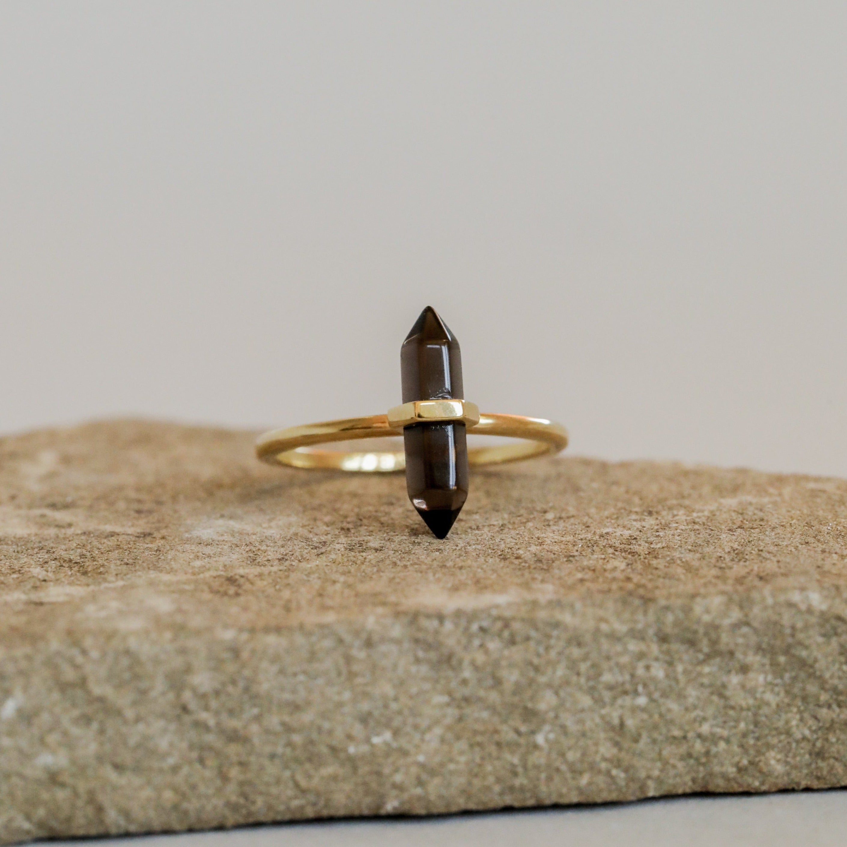 The Smoky Quartz Point Ring, featuring a double-pointed dark crystal set horizontally on a gold band, rests on a rough beige stone with a plain light background.