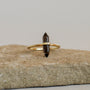The Smoky Quartz Point Ring, featuring a double-pointed dark crystal set horizontally on a gold band, rests on a rough beige stone with a plain light background.