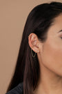 Close-up of a woman's ear adorned with the Stud Chain Huggie—an 18k gold plated earring featuring a small hoop with chain, perfect for stacking with other studs and cuffs. She has straight, dark brown hair against a light beige backdrop.