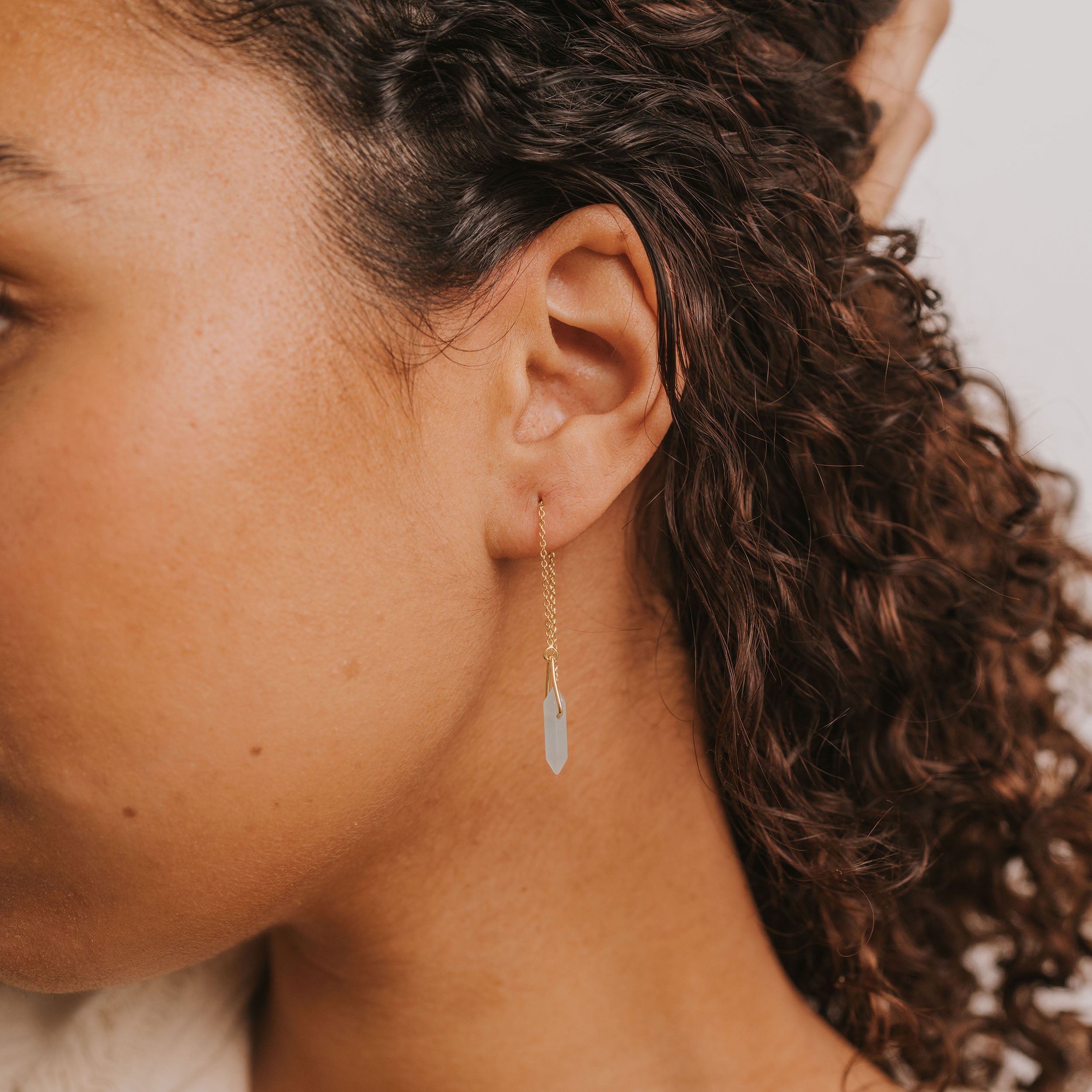 Close-up of a person with curly brown hair wearing the JaxKelly Gemstone Threader - Amazonite, a gold plated threader earring featuring a light blue amazonite gemstone pendant. Set against a plain white background to highlight this lasting piece.