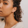 A close-up of a woman with curly hair wearing JaxKelly's Gemstone Threader - Amazonite gold plated earrings with a geometric charm. She is dressed in a textured cream top, looking slightly away from the camera against a plain background.