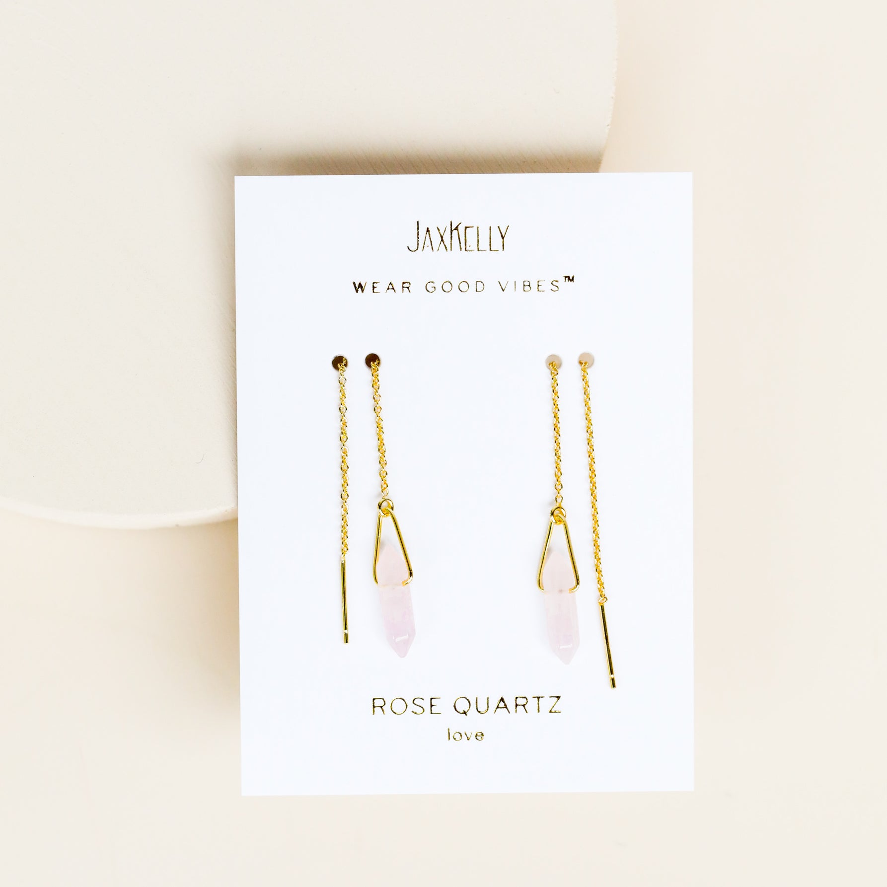 A pair of JaxKelly Gemstone Threader - Rose Quartz earrings with rose quartz crystals, gold plated, displayed on a white card that says "Wear Good Vibes - Rose Quartz, love." Background is soft beige.