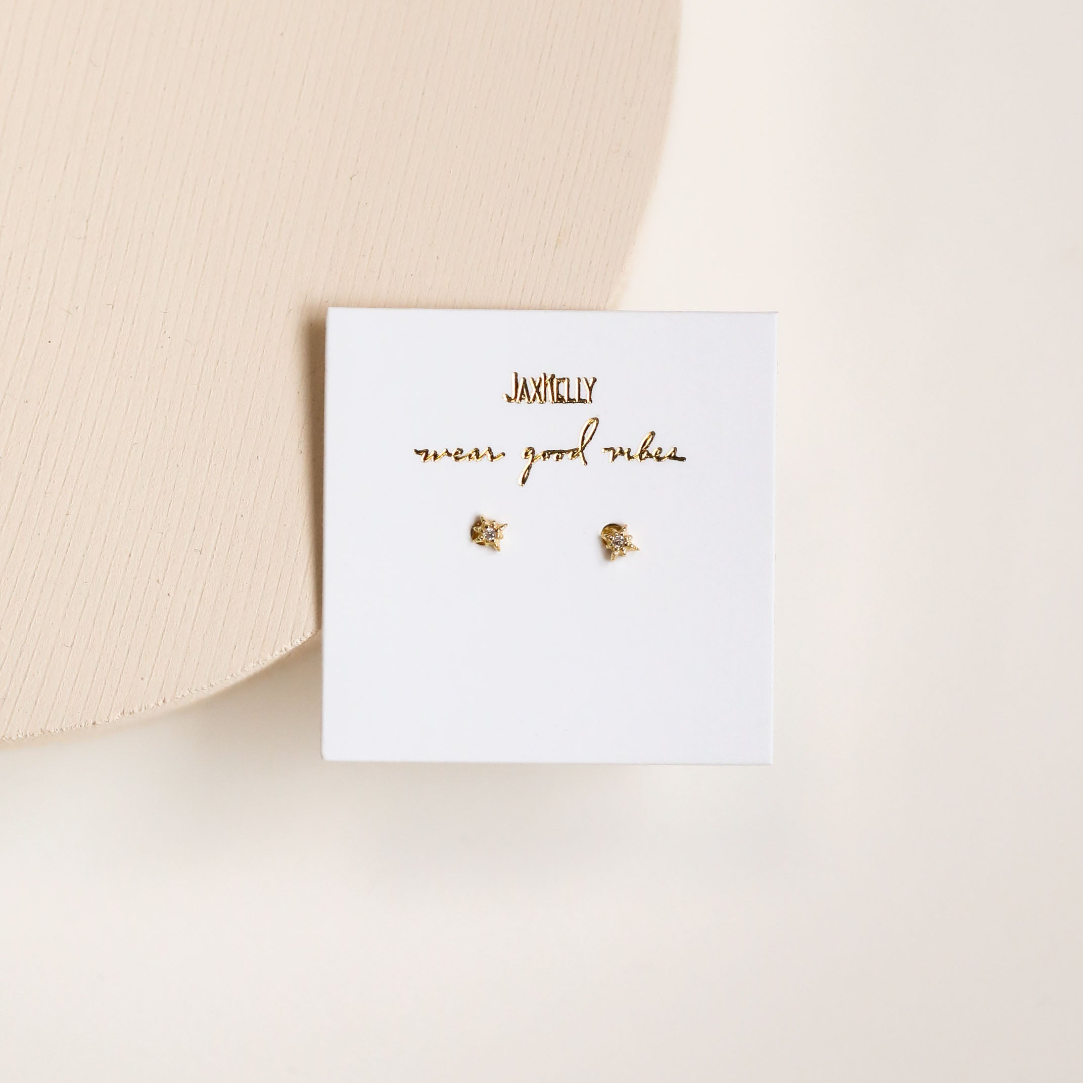 The Tiny Star mini stud earrings feature sterling silver posts and an 18k gold-plated finish, displayed on a white card with gold "JAXKELLY" and "wear good vibes" text, set against a neutral background.