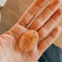 A hand with two gold rings holds the Citrine Touch Stone, a polished, oval-shaped natural crystal. The background features a brown wooden floor and part of a woven mat.