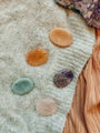 The Citrine Touch Stone is shown with three other polished gemstone touchstones in orange, green, and purple on soft knit fabric, next to amethyst crystals and a tan textured cloth.