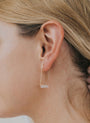 Close-up of a blonde person wearing the Rose Quartz Gemstone Drop - Love earring, featuring a minimalist 18k gold-plated design with a rectangular white stone hanging at the bottom, hair tucked to reveal the jewelry.
