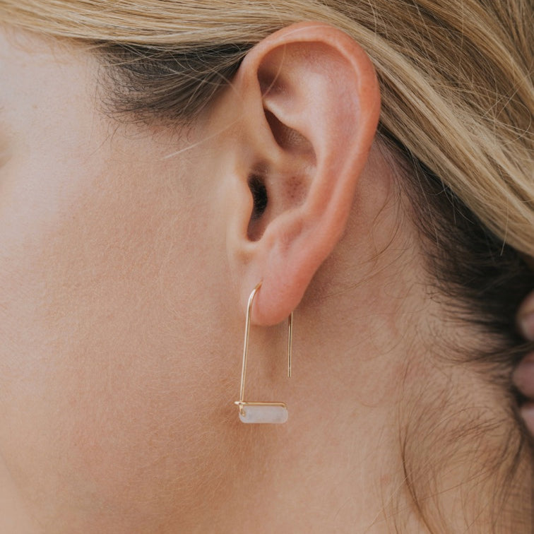 Close-up of a blonde person wearing the Rose Quartz Gemstone Drop - Love earring, featuring a minimalist 18k gold-plated design with a rectangular white stone hanging at the bottom, hair tucked to reveal the jewelry.