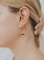 Close-up of a person with blonde hair wearing the Carnelian Agate Gemstone Drop - Creativity earring, featuring a minimalist gold-plated design with a horizontal rectangular reddish-brown stone, their hand highlighting the refined drop style.