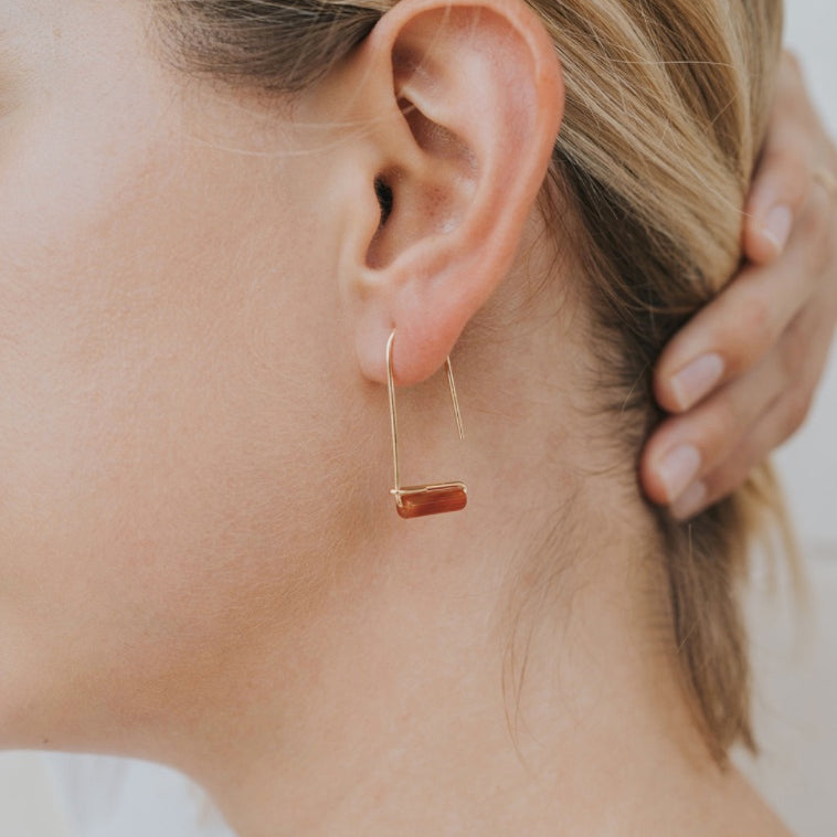 Close-up of a person with blonde hair wearing the Carnelian Agate Gemstone Drop - Creativity earring, featuring a minimalist gold-plated design with a horizontal rectangular reddish-brown stone, their hand highlighting the refined drop style.