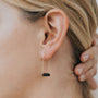 Close-up of a person with light skin and blonde hair wearing the Black Onyx Gemstone Drop - Focus: elegant gold plated drop earrings featuring a small horizontal black onyx bar, as they touch the back of their head.