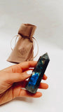 A hand holds a polished Labradorite Crystal Point, showing blue and green flashes. In the background, a tan JaxKelly drawstring pouch sits on a white surface.