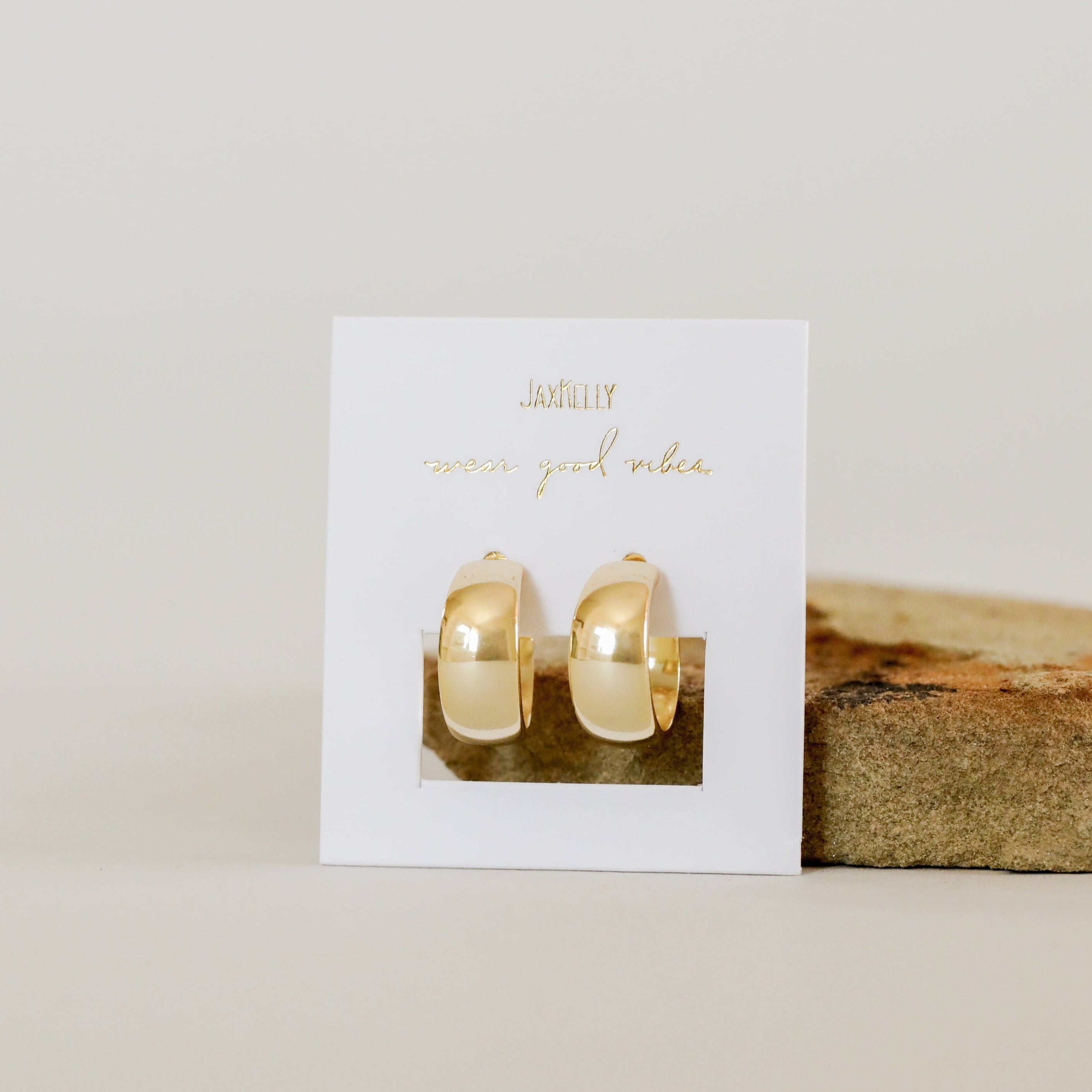 The Gold Wide Hoop earrings feature a shiny 18k gold-plated finish, displayed on a white card with gold text, set against a neutral background with a beige stone beside them.
