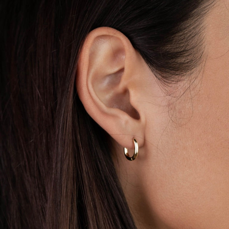 Close-up of a woman's ear with straight brown hair, wearing the Small Rectangular Hoop earring. The shiny gold hoop has sterling silver posts. Her skin looks smooth and the softly blurred background adds a gentle touch.