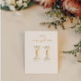 The Dual Triangle Drop earrings feature gold plating, crystal triangular drops, and sterling silver posts, displayed on a JaxKelly card with delicate flowers on a beige surface.