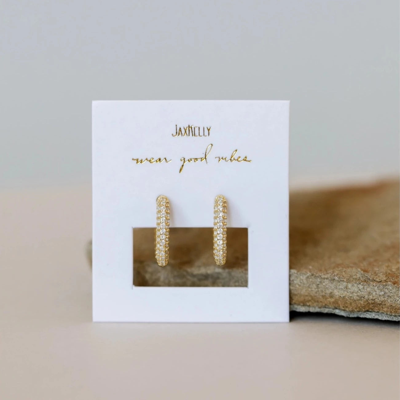 The Pavé Gold Hoop features 18k gold plated square hoops adorned with sparkling pave CZ stones, shown on a white “JaxKelly” card with “wear good vibes” gold text, set atop a neutral background.
