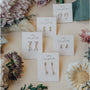 Five pairs of Dual Triangle Drop CZ earrings with gold-plated and sterling silver posts are displayed on white “Jaxlily wear good vibes” cards, surrounded by soft-colored flowers and greenery on a light surface.