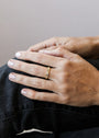 A close-up of a hand with neatly manicured nails and light polish, wearing dark pants, showcases the Inset Baguette Ring—a gold stacking ring featuring a small rectangular gemstone.