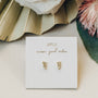 A pair of Offset Trio - White CZ stud earrings with delicate 18k gold plating rest on a white JaxKelly card, surrounded by neutral dried flowers and green leaves.