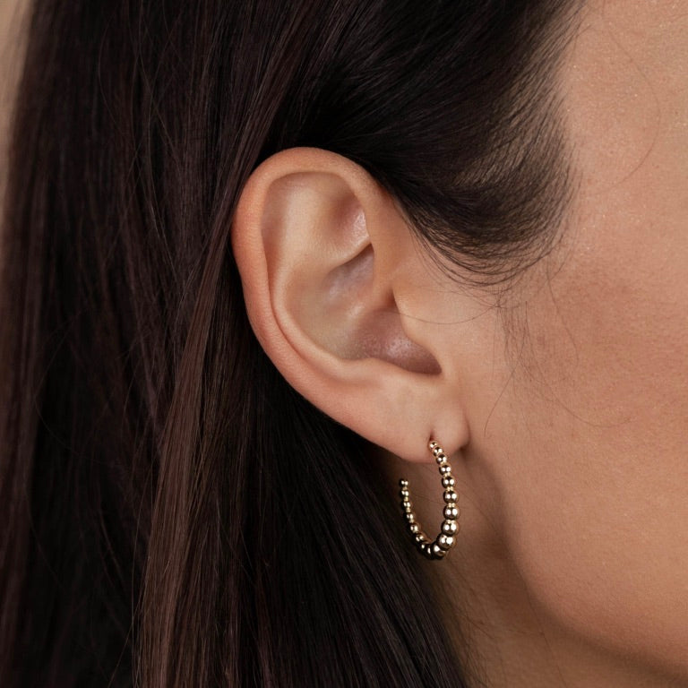 A close-up of a woman's ear with straight dark brown hair, wearing the Large Ball Hoop— a gold plated earring featuring a beaded texture and sterling silver posts.