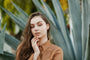A young woman with long brown hair and a tan blouse poses outdoors before blue-green agave leaves, her hand lightly on her chin, showcasing her chic Pavé Silver Hoop earrings.
