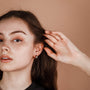 A young woman with long brown hair tucks it behind her ear, revealing the Silver Wide Hoop earring with minimalist design and sterling silver posts, as she gazes to the side against a plain beige background.