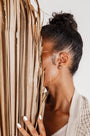 A woman with dark curly hair in a bun holds dried palm leaves, partially covering her face. She wears Silver Swirl Hoop earrings, a beige knit sweater, and has white-painted nails against a light background.