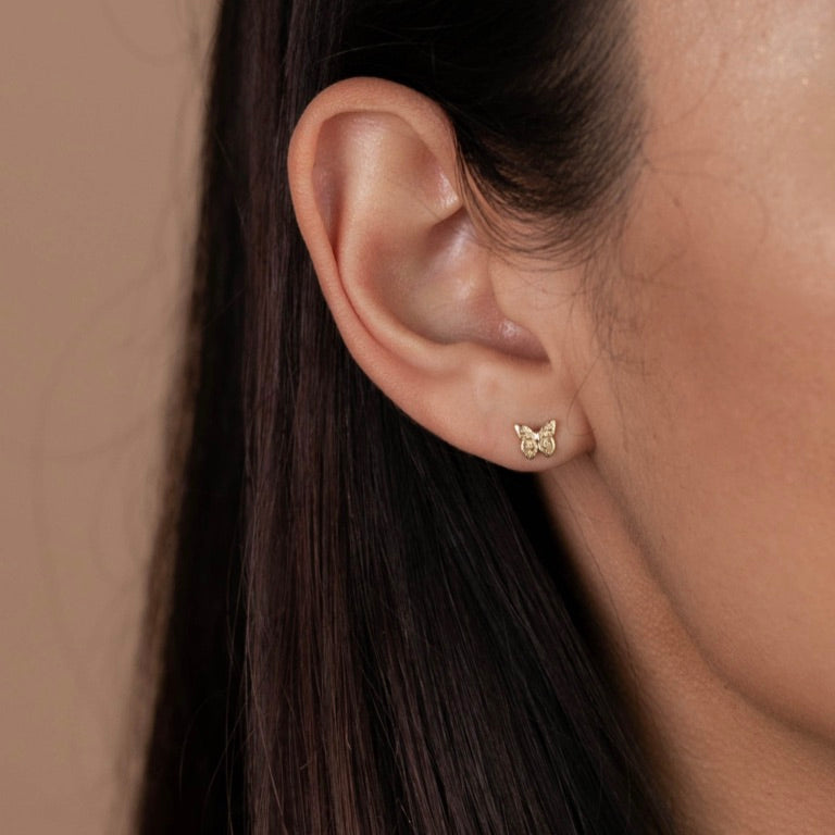 Close-up of a woman's ear with straight dark brown hair, wearing the Butterfly gold-plated stud earring with sterling silver posts against a beige background.