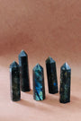Five Labradorite Crystal Points with iridescent blue-green flashes stand upright on a light brown surface, each radiating a protective aura, arranged in a loose row against a plain brown background.
